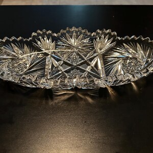 May include: A clear, cut-glass serving dish with three compartments. The dish has a scalloped edge and intricate starburst and geometric patterns. The glass reflects light, creating a sparkling effect. This decorative piece is suitable for serving snacks or appetizers.