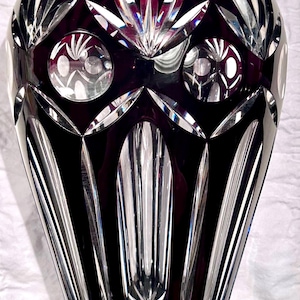 May include: A tall, elegant, dark red crystal vase with intricate cut patterns. The vase features a series of vertical lines, diamond shapes, and circular designs. The crystal reflects light, creating a sparkling effect. A decorative piece for home decor.