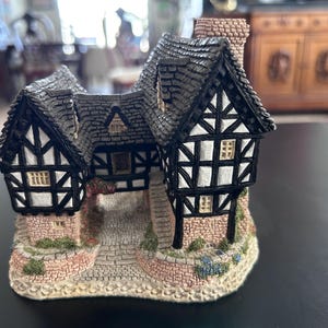 May include: A miniature Tudor-style cottage figurine. The house features a black and white timber-framed design with a gray shingled roof and a brick chimney. The base is made of brick and stone, with small floral accents.