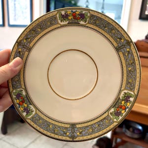 May include: A cream-colored saucer with a gold rim and a decorative border featuring a repeating pattern of blue, yellow, and gold scrollwork. The border includes images of fruit baskets. A gold ring encircles the center.