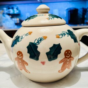 May include: A white ceramic teapot with a green and brown gingerbread man design. The teapot has a lid and a handle.