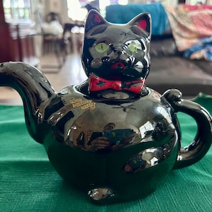 May include: A black ceramic teapot shaped like a cat with green eyes and a red bow tie. The teapot has a glossy finish and a handle on the side. The text "From Quebec Canada" is on the side.