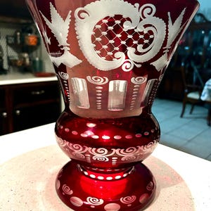 May include: A ruby red glass vase with intricate white etched designs. The vase features a flared top, a bulbous midsection, and a circular base. The etched patterns include floral and geometric motifs, adding a touch of elegance to the decorative piece.
