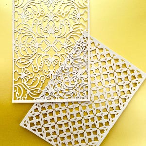 K&ouml;nnte beinhalten: Zwei wei&szlig;e Papierservietten mit filigranen Ausschnitten. Die obere Serviette hat ein Blumenmuster, w&auml;hrend die untere Serviette ein geometrisches Muster hat.