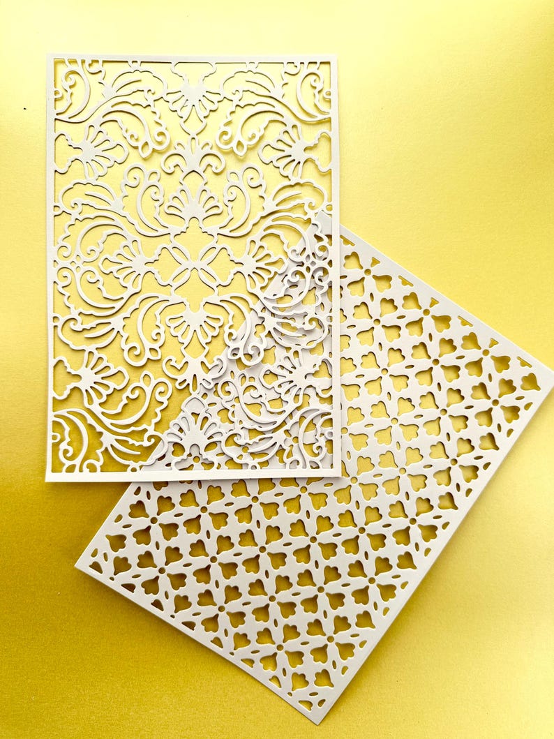 K&ouml;nnte beinhalten: Zwei wei&szlig;e Papierservietten mit filigranen Ausschnitten. Die obere Serviette hat ein Blumenmuster, w&auml;hrend die untere Serviette ein geometrisches Muster hat.