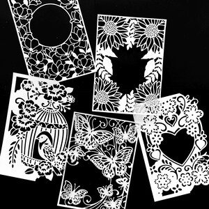 Può includere: Cinque disegni di carta ritagliati in bianco con intricati motivi floreali e di farfalle. I disegni sono disposti in uno schema casuale su uno sfondo nero.