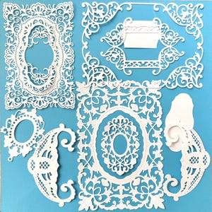 May include: A selection of white, intricately cut paper embellishments, featuring ornate frame and border designs. The collection includes oval and rectangular frames, decorative corners, and flourishes, set against a light blue backdrop. Suitable for scrapbooking and paper crafts.