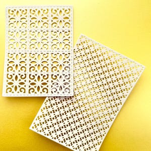 K&ouml;nnte beinhalten: Zwei wei&szlig;e dekorative Papierschablonen mit komplizierten geometrischen Mustern. Die Schablone links hat ein sich wiederholendes quadratisches Muster mit einem floralen Design. Die Schablone rechts hat ein sich wiederholendes kreisf&ouml;rmiges Muster.