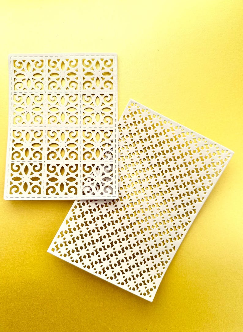K&ouml;nnte beinhalten: Zwei wei&szlig;e dekorative Papierschablonen mit komplizierten geometrischen Mustern. Die Schablone links hat ein sich wiederholendes quadratisches Muster mit einem floralen Design. Die Schablone rechts hat ein sich wiederholendes kreisf&ouml;rmiges Muster.