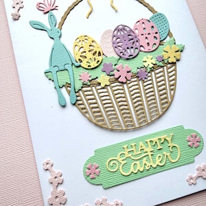 May include: An Easter card with a decorative basket filled with colourful eggs and flowers. A light blue bunny sits on the side of the basket. The words "Happy Easter" are displayed on a green banner.