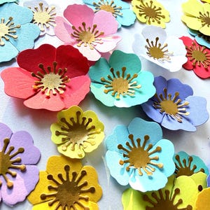 Könnte beinhalten: Eine Sammlung mehrfarbiger Papierblumen mit fünf abgerundeten Blütenblättern in Rot, Rosa, Gelb, Blau und Violett. Jede Blume hat in der Mitte ein metallisch goldenes Stanzteil mit dünnen, strahlenförmigen Linien als Staubgefäße.