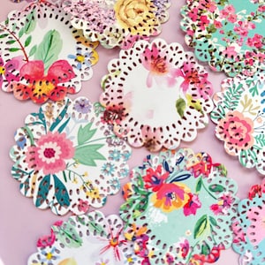 Può includere: Una collezione di centrini di carta floreali con bordi smerlati. Ogni centrino presenta un design floreale ad acquerello unico nei toni del rosa, giallo, blu e verde. I centrini sono disposti su una superficie rosa chiaro.