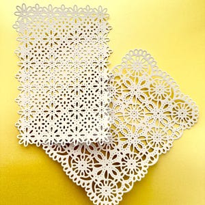K&ouml;nnte beinhalten: Zwei wei&szlig;e Papierdeckchen mit filigranen Blumenmustern. Die Deckchen liegen &uuml;bereinander, das kleinere Deckchen oben. Die Deckchen liegen auf einem gelben Hintergrund.