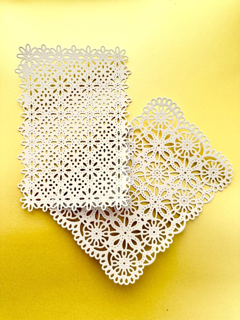 K&ouml;nnte beinhalten: Zwei wei&szlig;e Papierdeckchen mit filigranen Blumenmustern. Die Deckchen liegen &uuml;bereinander, das kleinere Deckchen oben. Die Deckchen liegen auf einem gelben Hintergrund.