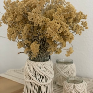 Könnte beinhalten: Drei Glasvasen mit weißen Makramee-Bezügen. Die Vasen stehen auf einer Holzoberfläche mit einem weißen Häkel-Läufer. Die Vasen sind mit getrockneten Blumen gefüllt.