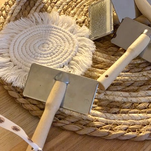 Könnte beinhalten: Drei Bürsten mit Holzgriff und Metallborsten auf einer weißen gewebten Matte mit einem braunen Tontopf im Hintergrund.