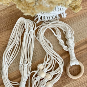 Könnte beinhalten: Ein weißer Makramee-Pflanzenhänger mit Holzperlen und einem Holzring. Der Hänger ist aus Baumwollschnur gefertigt und hat ein geknüpftes Design.