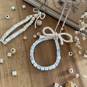 Könnte beinhalten: Zwei weiße Buchstabenperlen-Lesezeichen, eines mit einem silbernen Büroklammer und einer weißen Schleife, das andere mit einem silbernen Büroklammer und einer Holzperle. Die Perlen liegen auf einer Holzoberfläche.