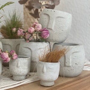 Könnte beinhalten: Ein Set aus vier grauen Beton-Pflanzgefäßen in Form von Gesichtern. Die Gefäße sind in verschiedenen Größen und auf einer weißen Oberfläche angeordnet. Die Gefäße sind mit einer strukturierten Oberfläche verziert und haben eine dezente graue Farbe.