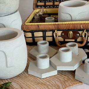 Könnte beinhalten: Ein Set aus fünf grauen Beton-Kerzenhaltern mit sechseckigen Sockeln. Die Kerzenhalter sind auf einer gewebten braunen Matte angeordnet. Im Hintergrund befindet sich ein Weidenkorb mit einem braunen Ledergriff.