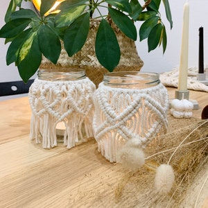 Könnte beinhalten: Zwei Glas-Kerzenhalter mit weißen Makramee-Bezügen. Die Kerzenhalter stehen auf einem Holztisch mit getrockneten Blumen und einer Pflanze in einem geflochtenen Korb.