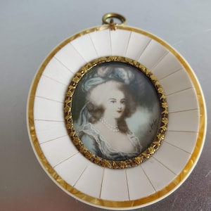 Op de afbeelding: Een ronde ivoor en gouden lijst met een miniatuurportret van een vrouw in een blauwe en witte jurk. De vrouw draagt een parelketting en heeft haar haar in een gepoederde pruik gestyled. De lijst heeft een gouden rand en een kleine lus aan de bovenkant om op te hangen.