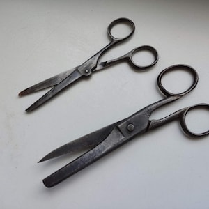May include: Two pairs of vintage scissors with metal handles. The larger pair has a longer blade and a more rounded handle. The smaller pair has a shorter blade and a more pointed handle.