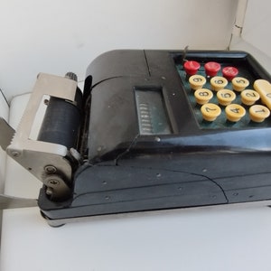 Op de afbeelding: Een zwarte vintage kassa met een papierrolhouder, een displayvenster en een numeriek toetsenbord met rode en gele knoppen. De kassa heeft een scheur aan de zijkant.