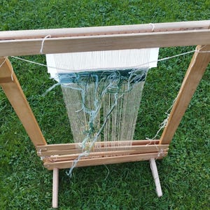 Può includere: Un telaio in legno con fili di ordito chiari tesi verticalmente. Un tessuto parzialmente lavorato con filato verde scuro e azzurro è fissato all'ordito. Il telaio presenta una base robusta e una barra superiore con viti metalliche a vista.