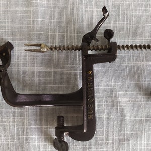 Puede incluir: Un pelador de manzanas vintage de hierro fundido negro con un mango de madera y una abrazadera de resorte. El pelador está etiquetado como "HUEY CHERRY".