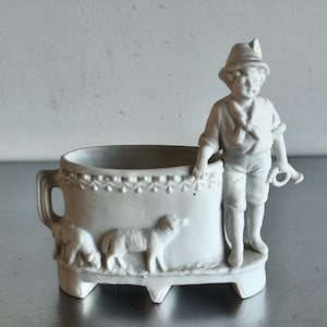 Può includere: Un vaso di porcellana bianca con manico e tre piedi. Il vaso presenta un rilievo di un pastore con un cappello, che tiene in mano uno strumento e due pecore. Il bordo del vaso ha un motivo decorativo.