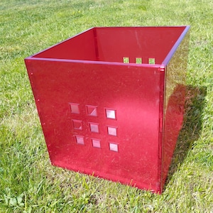 May include: A translucent red square storage bin constructed from rigid acrylic panels. The front panel features a grid of nine small, square cutouts arranged in a three-by-three pattern, while the top rim remains open for access.