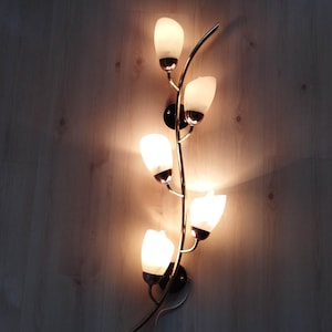 Può includere: Una lampada da parete con uno stelo metallico curvo che si ramifica per sostenere cinque paralumi individuali in vetro smerigliato a forma di tulipano. Ogni paralume emette una luce calda e luminosa, creando una disposizione verticale.