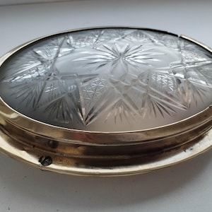 May include: An antique, round ceiling light fixture with a frosted glass shade featuring a starburst and geometric pattern. The light has a brass-colored metal frame and a black wire.