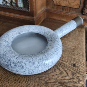 Op de afbeelding: Een rond, grijs en wit gespikkeld keramisch object met een handvat. Het object heeft een cirkelvormige opening in het midden en een metalen dop op het handvat. Het item ligt op een houten oppervlak.