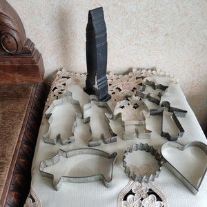 Peut inclure: Un ensemble de découpoirs à biscuits en métal vintage de différentes formes, notamment des personnes, un cochon, un cœur et une couronne. Les découpoirs sont disposés sur une nappe blanche avec un découpoir à biscuits en métal noir en arrière-plan.