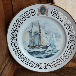 Op de afbeelding: Witte porseleinen plaat met een gouden rand en een zwarte en witte decoratieve rand. Het midden van de plaat toont een schilderij van een schip dat op de oceaan vaart met ijsbergen op de achtergrond.