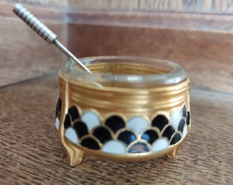 May include: A small, gold-colored salt cellar with a clear glass top and a black and white geometric design. The cellar has three legs and comes with a small, silver-colored spoon.