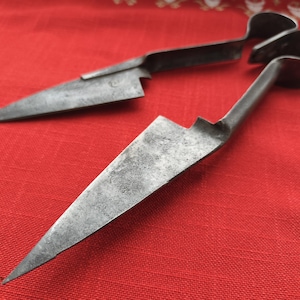 May include: Two antique, metal shears with sharp, triangular blades and curved handles. The shears are a dark, aged metal, resting on a red fabric surface. The blades are angled away from each other, suggesting they are open and ready for use.