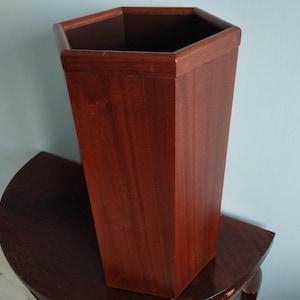 Könnte beinhalten: Ein hoher, sechseckiger Holzbehälter mit einer satten, rotbraunen Oberfläche. Der Behälter hat ein geometrisches Design und steht auf einem dunklen, halbkreisförmigen Tisch. Der Innenraum ist dunkler braun.