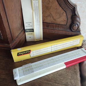 May include: A yellow and red slide rule with the word "ARISTO" printed on the side. The slide rule is on a wooden surface. There is a smaller instruction booklet for the slide rule with the words "ARISTO JUNIOR" printed on the front.
