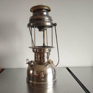 May include: A vintage-style metal pressure lantern featuring a polished silver-tone finish, a rounded fuel tank base, a central burner assembly, and a brass-coloured top cap. The lantern includes a vertical handle and small adjustment knobs.