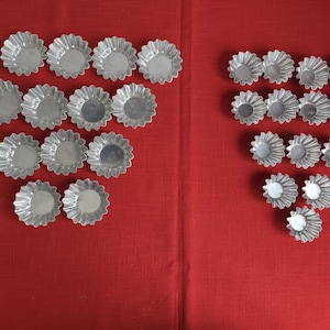 Puede incluir: Vista cenital de una colección de moldes para tartaletas plateados y acanalados sobre un mantel rojo. Los moldes, de diferentes tamaños, están dispuestos en dos grupos. Tienen un borde festoneado y una base circular, ideales para repostería.