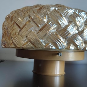 Könnte beinhalten: Eine Vintage-Leuchte mit einem gewebten Glasschirm. Der Schirm hat eine hellbraune Farbe und sitzt auf einem beigefarbenen Metallsockel. Die Leuchte ist für die Deckenmontage konzipiert.