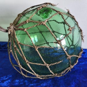 Op de afbeelding: Een grote, ronde, groene glazen visdrijver, omhuld door een bruin touwnet. De drijver heeft een metalen bevestiging en is gedeeltelijk gevuld met een donkerblauwe substantie. Het glas heeft een lichte textuur en een klein groen glazen stukje bovenop.