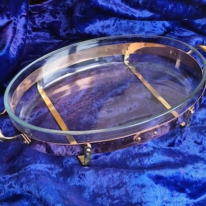 Könnte beinhalten: Eine ovale Glasbackform mit einem kupferfarbenen Metallrahmen und Griffen. Die Form wird von einem goldfarbenen Metallständer getragen. Der Hintergrund ist ein tiefblauer Samtstoff.