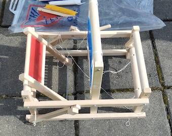 May include: A wooden loom with a red and white woven piece on the loom. The loom has a blue and white string attached to it.