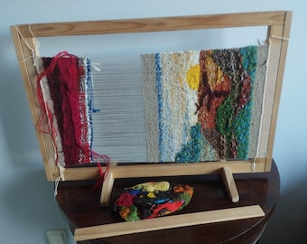 May include: A wooden weaving loom with a partially completed tapestry. The tapestry features red, blue, and white threads on the left, transitioning to a colorful abstract design on the right. A small bundle of colorful yarn sits below.