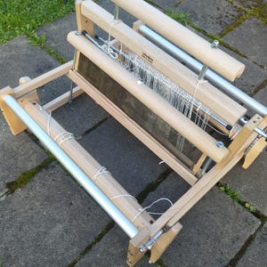 Puede incluir: Un telar de mesa de madera clara con un peine de metal, hilos de urdimbre blancos y dos rodillos cilíndricos. El marco está construido con madera lisa sin acabado, herrajes de metal plateado y una perilla de ajuste negra en el lateral.