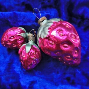 May include: Three glass strawberry ornaments with pink and red coloring and green accents. The ornaments have a metallic finish and are arranged on a blue velvet surface. Each ornament has a metal loop for hanging.